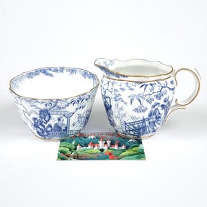 Könnte beinhalten: Ein blau-weißes Zuckerdosen- und Sahnekännchen-Set. Das Set verfügt über ein detailliertes blaues Blumen- und Landschaftsdesign mit goldenen Zierleisten. Das Sahnekännchen hat einen Griff und einen Ausguss. Die Zuckerdose ist rund. Eine kleine Karte liegt vor dem Set.