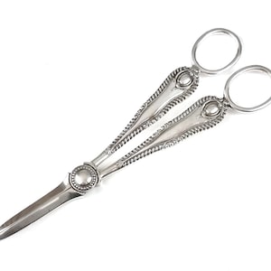 May include: Silver-plated scissors with ornate handles and a pointed blade. The handles feature a decorative design and a circular detail at the centre.
