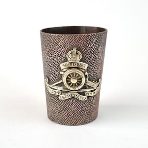 May include: A copper-coloured metal cup with a textured surface. The cup features a gold-coloured emblem with a crown, a cannon, and the words "Ubique et Gloria Ducis".