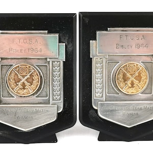 Puede incluir: Dos placas plateadas con respaldo negro, cada una con un diseño de diana dorada y rifles cruzados. La placa superior dice "F.T.C.S.A. Bisley 1964" y la placa inferior dice "S/L M/G Team Match Winners". La segunda placa dice "F.T.C.S.A. Bisley 1964" y la placa inferior dice "S/L Revolver Team Match Winners".