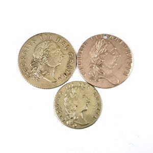 May include: Three gold-colored coins with a profile of a person and Latin text around the edge. The coins are of different sizes, with the largest coin at the top and the smallest at the bottom. The coins are on a white background.
