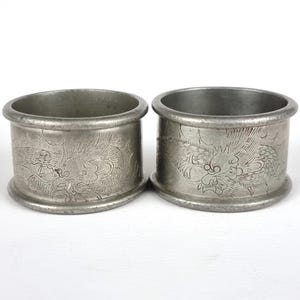 May include: Two antique silver napkin rings. Each ring features an embossed dragon design. The rings are cylindrical with a slightly flared base and top. The metal has a matte finish and shows signs of age.