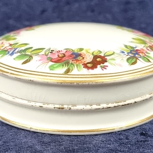 Puede incluir: Caja de porcelana blanca para alhajas con borde dorado y diseño floral. La tapa está decorada con una corona de flores de colores.