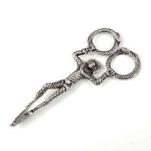 May include: A pair of silver-toned metal scissors featuring a decorative handle shaped like a figure wearing a wide-brimmed hat and a ruffled collar. The finger loops are textured to resemble coiled snakes, and the blades form the figure's legs.