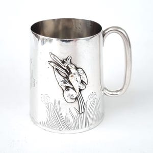 Puede incluir: Taza de plata con un diseño en relieve de pájaros y juncos. La taza tiene un asa redondeada.