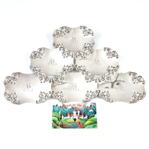 Pode incluir: Um conjunto de seis pratos de servir decorativos prateados. Cada prato tem uma borda ondulada com ornamentos ornamentais e um monograma central. Um cartão colorido com uma cena de castelo está em primeiro plano.