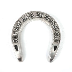 May include: A silver-toned metal horseshoe with a smooth, polished finish. The surface features embossed capital lettering that reads "GEH AUF BATA DU FAHRST GUT" along the curved top edge, with two small triangular cutouts near the bottom ends.