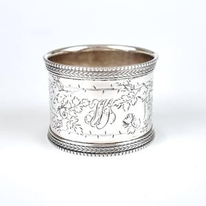 May include: Silver napkin ring with an engraved floral design and a monogram. The ring is circular and has a beaded edge.
