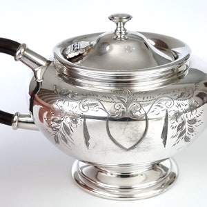 May include: A silver teapot with an ornate engraved design and a black wooden handle. The teapot has a lid with a finial.