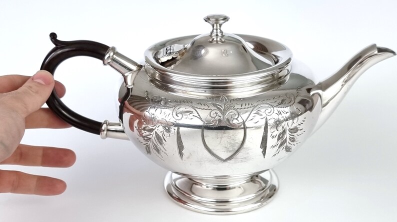 May include: A silver teapot with an ornate engraved design and a black wooden handle. The teapot has a lid with a finial.