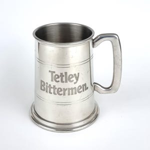 May include: A stainless steel beer mug with a handle. The mug has horizontal bands and the words "Tetley Bittermen" are etched on the front. The mug is designed for holding beer or other beverages.
