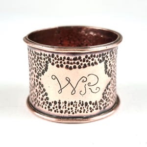 May include: A copper napkin ring with a dotted texture. It has an engraved cartouche with the intertwined initials "WR". The metal has a warm, reddish-brown hue. The ring is approximately 4 cm in diameter.
