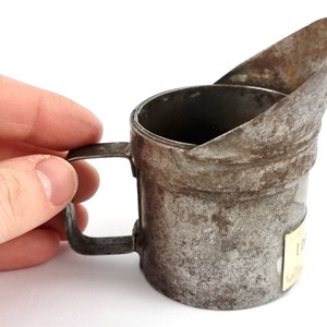 Pu&ograve; includere: Una vecchia lattina di olio in metallo con un beccuccio e un manico. La lattina &egrave; arrugginita e ha un'etichetta sul lato.