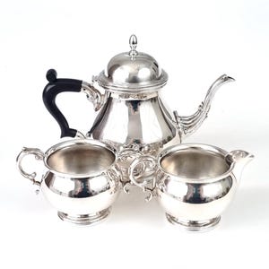 Puede incluir: Un juego de té plateado con tetera, jarra para la crema y azucarero. La tetera tiene un asa negra y una tapa con un pequeño pomo. El juego está muy pulido y refleja la luz, creando una apariencia brillante.