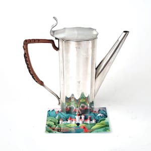Könnte beinhalten: Eine silberne Kaffeekanne mit langem Ausguss und geflochtenem braunen Griff. Die Kanne hat ein geriffeltes Design und einen dekorativen Deckel. Eine farbenfrohe Karte mit einer Schlosslandschaft befindet sich am Boden.