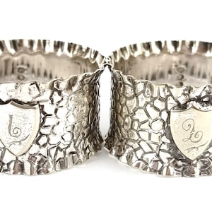 Op de afbeelding: Een paar zilveren servetringen met een getextureerd ontwerp en een wapenschild met de letter "L" op elke ring.