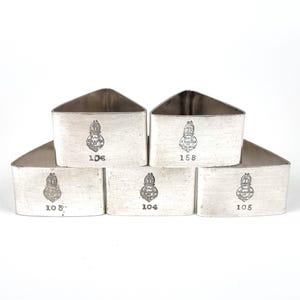 Könnte beinhalten: Fünf silberne Serviettenringe, jeweils mit einer eindeutigen Nummer und einem Wappenmotiv. Die Serviettenringe sind rechteckig mit einer dreieckigen Oberseite. Die Nummern sind schwarz gedruckt. Die Wappen sind detailliert.