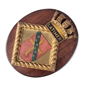 May include: A wooden plaque with a gold rope border and a crest featuring a red and green shield with a gold cross and fleur-de-lis. The plaque also has a gold crown and the text "LATIMER" in black.