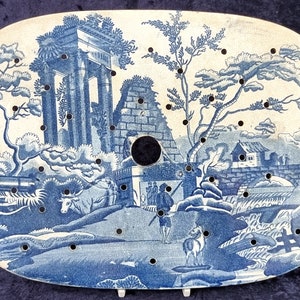Puede incluir: Un plato de cerámica azul y blanco con un diseño decorativo de un paisaje con un puente, árboles y un edificio. El plato tiene un borde festoneado y un agujero en el centro.