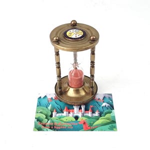 Puede incluir: Un reloj de arena de latón con un bulbo de vidrio central que contiene arena rosa. La parte superior presenta un emblema circular con un escudo de cuatro partes. Debajo, una tarjeta rectangular muestra una ilustración de un castillo blanco y un dragón rojo.