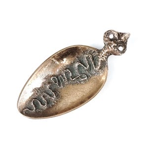 Può includere: Un piccolo cucchiaio dorato con un manico decorativo. Il cucchiaio ha la forma di una lacrima e una superficie testurizzata. Il manico ha la forma di una testa di uccello e ha un piccolo foro nella parte superiore.