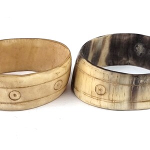 May include: Two carved bone bracelets with a decorative pattern of circles and lines. The bracelets are light brown and have a natural, rustic look.