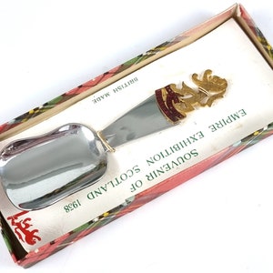 May include: Silver souvenir spoon with a gold-colored design of a lion and the text "SOUVENIR OF SCOTLAND EXHIBITION 1938 EMPIRE" on the handle. The spoon is in a red and green plaid box with the text "BRITISH MADE".