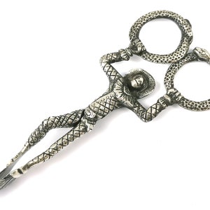 Può includere: Pinzette per lo zucchero argentate a forma di persona che tiene due serpenti in cerchio. La persona indossa un abito a motivi e un cappello.