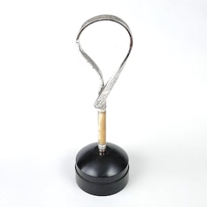 May include: A silver-toned metal loop with intricate engraved patterns attached to a cylindrical cream-coloured handle, which is mounted vertically onto a solid black circular base. The object stands upright with a smooth, polished finish.