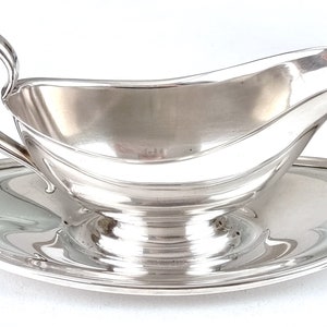May include: A silver gravy boat with a matching saucer. The boat has a rounded, curved shape with a handle on the left side. The saucer is round and has a slightly raised edge.
