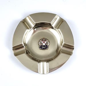 May include: A silver ashtray with a raised design of a coat of arms featuring a red cross on a white background, a blue banner with gold lettering, and a gold crown.