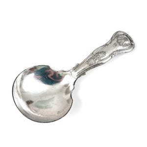 May include: A silver-toned metal spoon with a wide, rounded bowl and a short, ornate handle. The handle features embossed decorative patterns, including a shell-like motif at the tip and intricate scrollwork along the stem of the utensil.