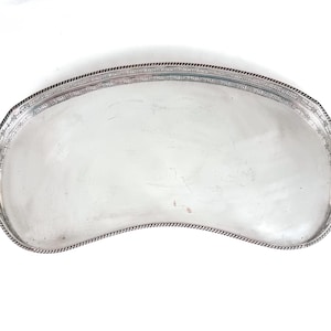 Puede incluir: Una bandeja de servicio plateada con forma de riñón que presenta una superficie pulida y reflectante, junto con un borde decorativo estilo cuerda a lo largo de todo el contorno. La bandeja tiene un diseño curvo y liso con un borde elevado.