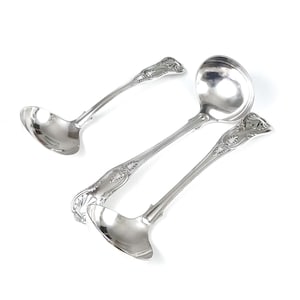 Può includere: Tre mestoli d'argento con dettagli floreali ornati sui manici. I mestoli sono disposti su uno sfondo bianco, con le conche rivolte in direzioni diverse. I cucchiai sono in metallo lucido e riflettente.