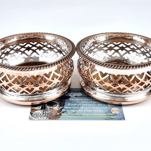 May include: Two silver and copper wine bottle coasters with a decorative openwork design. The coasters are round and have a raised rim.