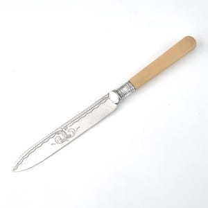 Könnte beinhalten: Ein silbernes Buttermesser mit einem Knochengriff. Die Klinge hat ein filigranes Design und der Griff ist hellcremefarben.