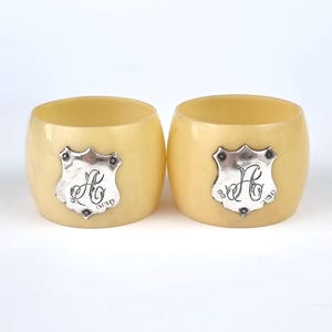 Puede incluir: Dos anillos de servilleta de marfil con escudos plateados grabados con las iniciales "H.C."