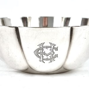 Puede incluir: Un cuenco de plata con un borde acanalado y un diseño de monograma. El monograma es un "HC" estilizado con un borde decorativo.