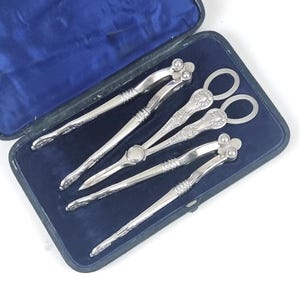 Puede incluir: Un juego de cascanueces y pinchos de plata antiguos, exhibidos en un estuche forrado de terciopelo azul. Los cascanueces tienen un diseño de palanca con patrones florales decorativos. Los pinchos tienen asas de bucle.