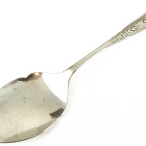 Puede incluir: Una cuchara de plata con un mango adornado y una letra "C" grabada en la parte superior. La cuchara tiene un cuenco ligeramente curvo y redondeado.