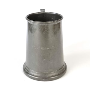 Könnte beinhalten: Ein Zinnhumpen mit Henkel und breitem Boden. Der Humpen hat eine leicht konische Form und einen kleinen Henkel. Die Oberfläche des Humpens hat ein mattes Finish und scheint mit dem Namen "A. Hamilton" graviert zu sein.