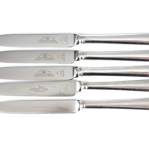 Puede incluir: Cinco cuchillos de mantequilla de plata con mango redondeado y hoja puntiaguda. Los cuchillos están grabados con el texto "A.E.W. & Co. Sheffield England Cutlers & Silversmiths" y "525/4"