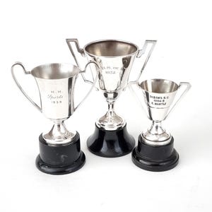 May include: Three silver trophies with black bases. The left trophy is inscribed "H.H. Sports 1939", the centre trophy is inscribed "B.S. of 1952", and the right trophy is inscribed "St. Bon's S.C. 1964-6 E. Mantle".