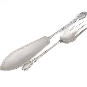 Può includere: Un set di coltello per torta e forchetta in argento con manici decorati e un design decorativo.