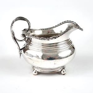 May include: A polished silver creamer featuring a rounded body with horizontal ribbed detailing, a decorative ornate handle, and a scalloped rim. The vessel stands on three small ball feet and includes a prominent pouring spout on the front side.