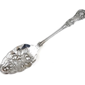 May include: A silver serving spoon with an ornate handle and a bowl decorated with a raised design of fruit and leaves.