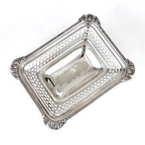 May include: A rectangular silver serving tray with ornate floral corner details. The tray has a perforated border with a repeating semi-circle pattern. The interior is a smooth, reflective surface. The tray is designed for serving food or displaying items.