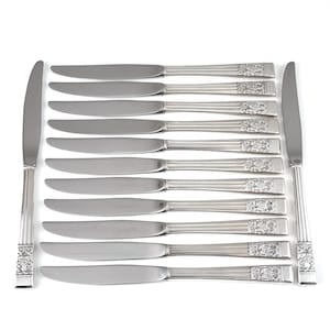Può includere: Un set di dodici coltelli da tavola in acciaio inossidabile. Le lame sono lisce e i manici presentano un motivo floreale decorativo. I coltelli sono disposti in file parallele, con un coltello su ogni lato.