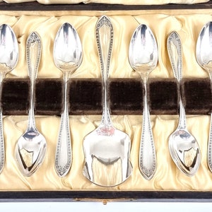 May include: A set of eight silver spoons in a wooden box with a velvet lining. The spoons are arranged in two rows of four, with a larger spoon in the centre. The spoons have ornate designs and are in excellent condition.