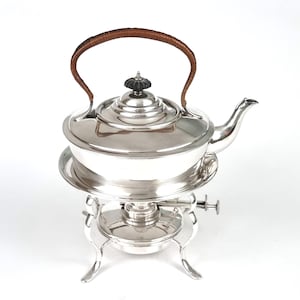 May include: A polished silver-plated tea kettle with a curved brown woven handle and a black finial on the lid. The kettle rests on a matching silver-plated stand featuring a burner mechanism with four curved legs and a side adjustment lever.